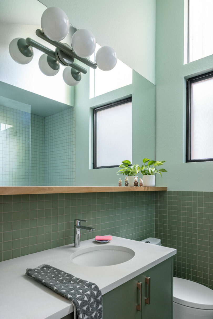 Modern bathroom with green tile walls, round sink on a white countertop, plant on the windowsill, three adjacent windows, and a light fixture with round bulbs above the mirror.