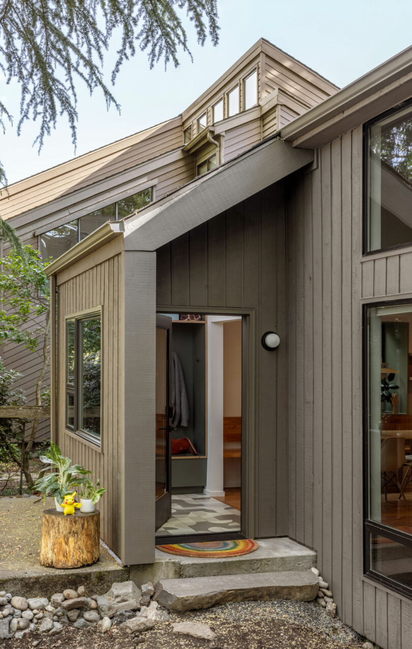 Modern house exterior with a geometric design, featuring wood paneling, a small porch with a round light fixture, and a colorful rug inside the entrance. Vegetation surrounds the house.