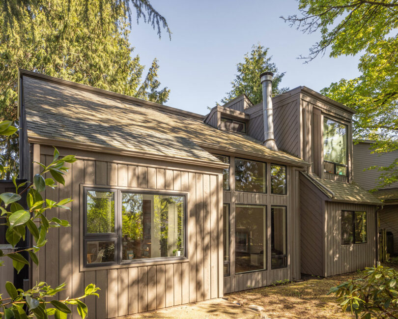 Modern two-story house with large windows, wood siding, and surrounded by trees under a clear sky.