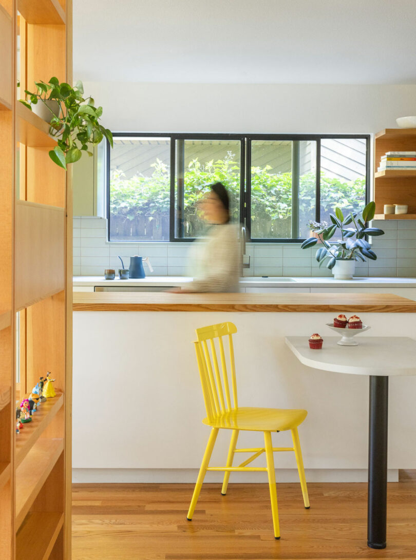 Modern kitchen with a yellow chair at a small table, cupcakes on the table, open shelf with decor, and a blurred person near the sink. Large windows with greenery in the background.