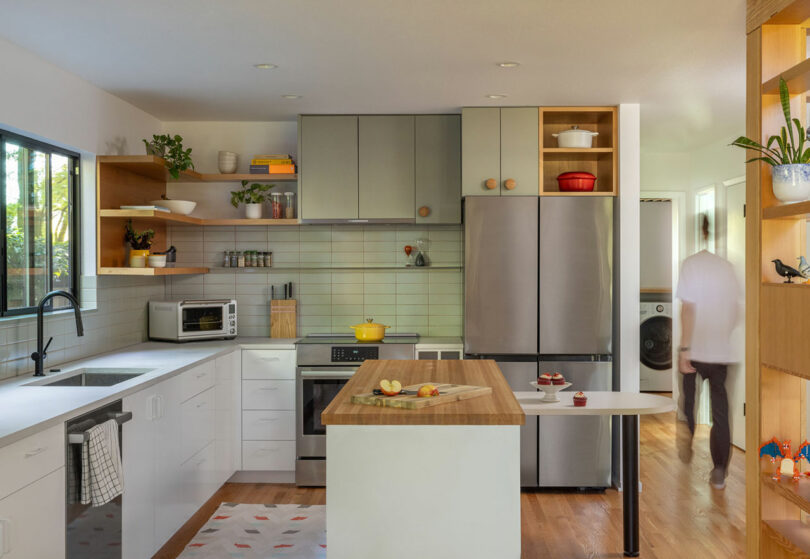 Modern kitchen with gray and white cabinets, stainless steel appliances, and a wooden island. A person is walking in the background near a laundry area.