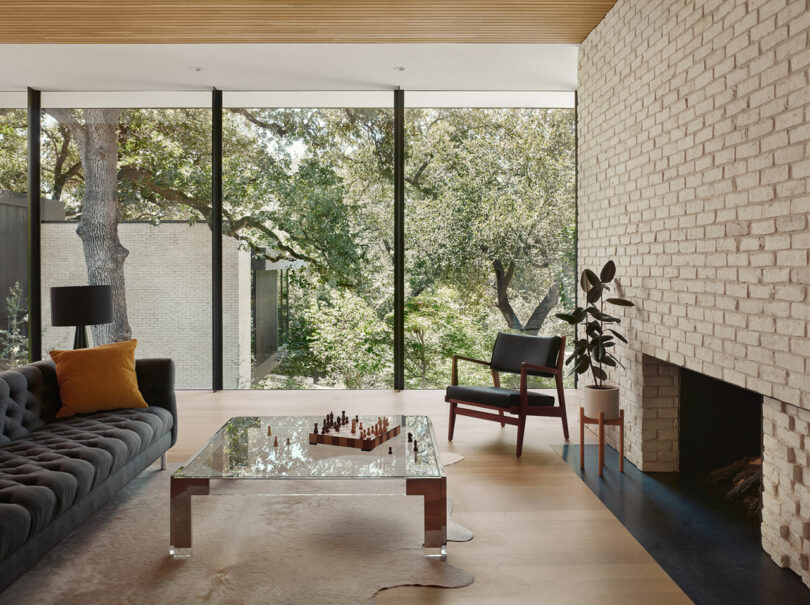 Modern living room with brick walls, large windows showing greenery, a glass coffee table with a chess set, a dark sofa with an orange pillow, a black chair, and a plant by the fireplace.