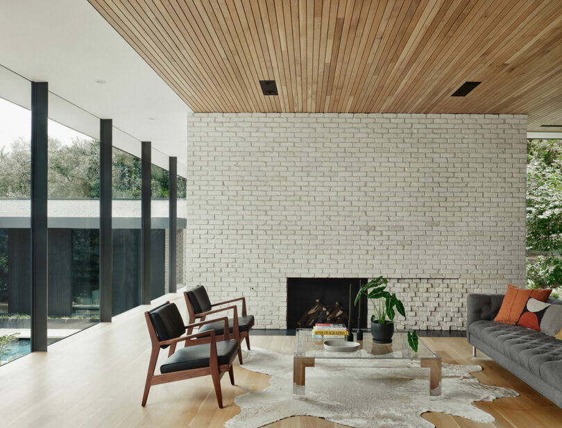 Modern living room with a white brick wall, fireplace, gray sofa, wooden chairs, and cowhide rug. Large windows reveal a view of trees.