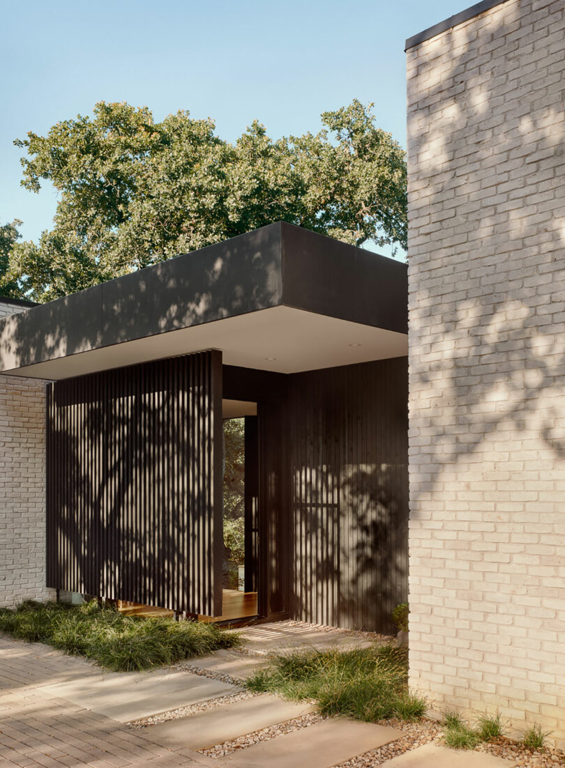 Modern house entrance with vertical wooden slats, white brick walls, and a flat roof. Shadows of trees visible on the walls.