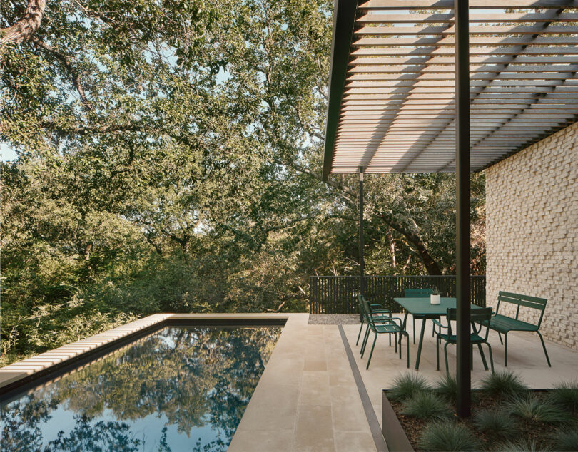 A modern outdoor patio features a rectangular pool, green table and chairs, and a pergola overhead, surrounded by lush trees.