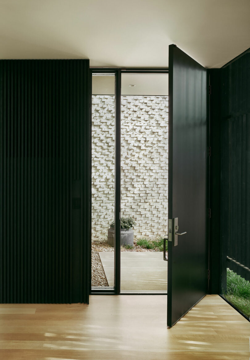 A modern, partially open black door with vertical grooves leads to a small courtyard with a brick wall and potted plant. Light wood flooring is visible inside.