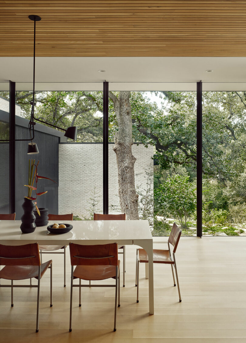 Modern dining area with a rectangular white table and five brown chairs. A black vase with branches sits on the table. Large windows reveal a view of trees and a white brick wall outside.