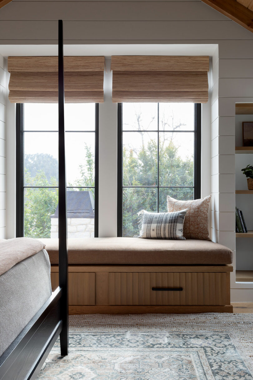 Bedroom with a black-framed bed, window seat with cushions, large windows, wooden blinds, and a patterned rug. Bookshelves are visible on the right.