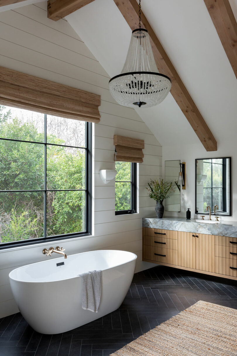 A modern bathroom with a freestanding bathtub, large window, wooden vanity with marble countertop, and a chandelier. Dark herringbone floor and natural light complete the scene.