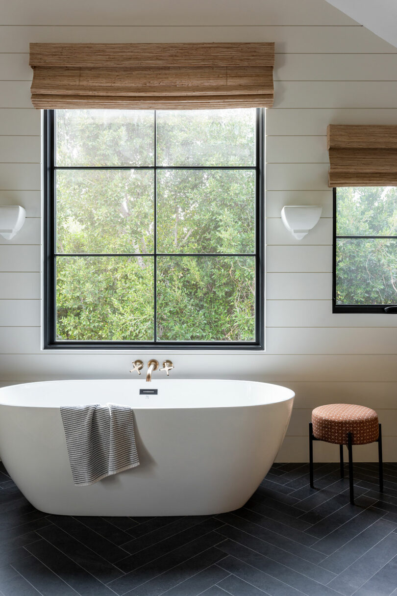 A modern bathroom with a white freestanding tub, a gray towel draped over the side, a window with a leafy view, a small stool, and two wall lights.