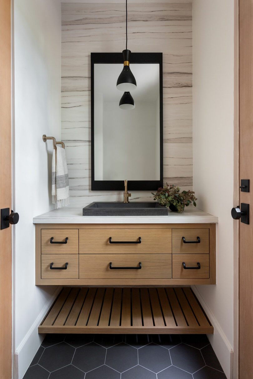 Modern bathroom with a wood vanity, black-framed mirror, black sink, and minimalist pendant light. White striped wall and hexagonal floor tiles add contrast. Towel on a brass holder.