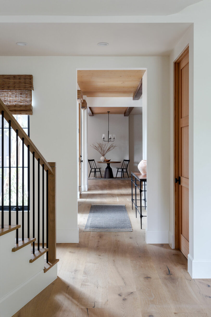 Interior hallway with wooden flooring, a staircase on the left, and a table with a vase and dried branches in the background. Two chairs and a chandelier are visible in the dining area.