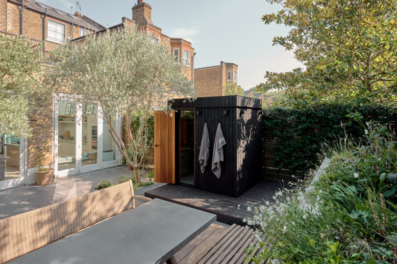 Outdoor garden shower with wooden privacy enclosure and hanging towels, surrounded by plants and trees, near a brick house with large windows and a patio table