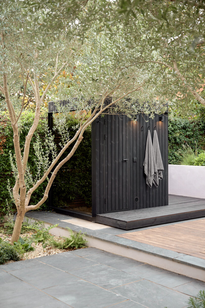 Outdoor wooden sauna with gray towels hanging, surrounded by greenery and a tile walkway in the foreground.