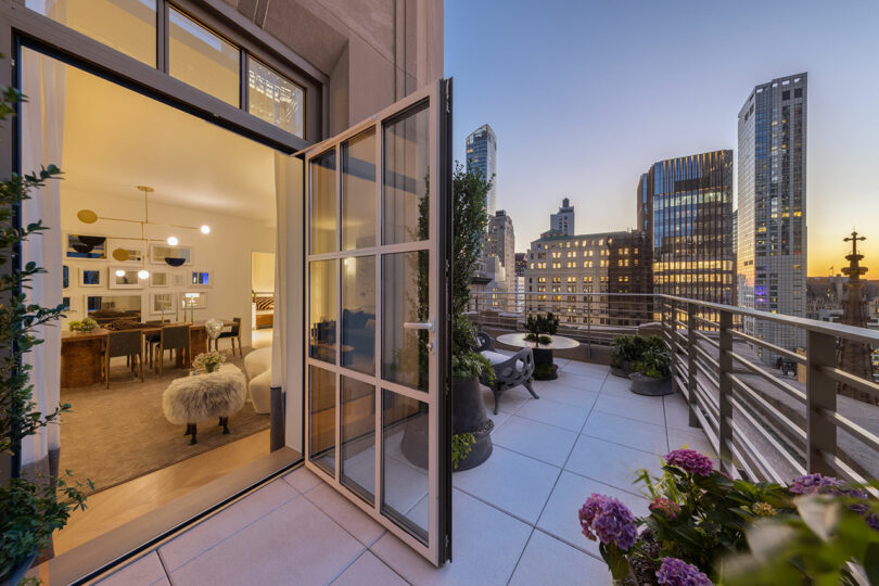 Open balcony door leading to a furnished apartment with dining area. Terrace, styled with a touch of Elizabeth Graziolo's elegance, features potted plants and offers a view of city buildings at sunset.