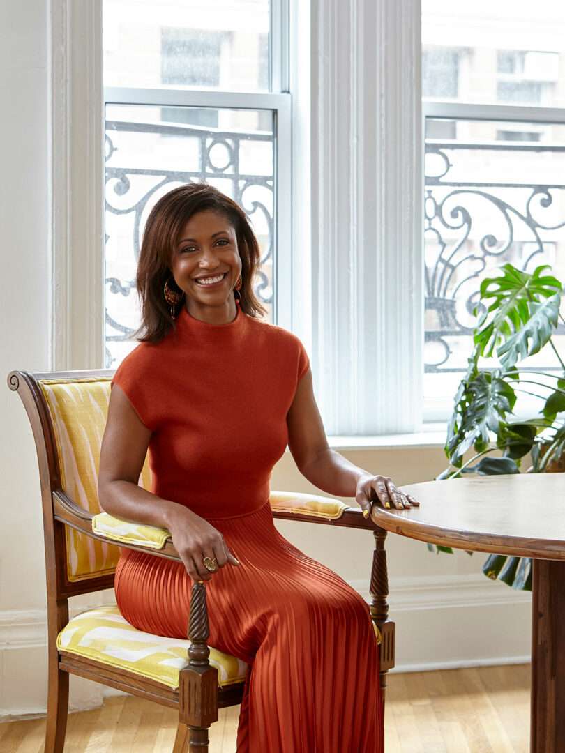 A person in an orange outfit sits on a patterned chair at a wooden table, basking in the sunlight streaming through large windows, reminiscent of a tranquil space designed by Elizabeth Graziolo, with a lush plant adding a touch of nature.