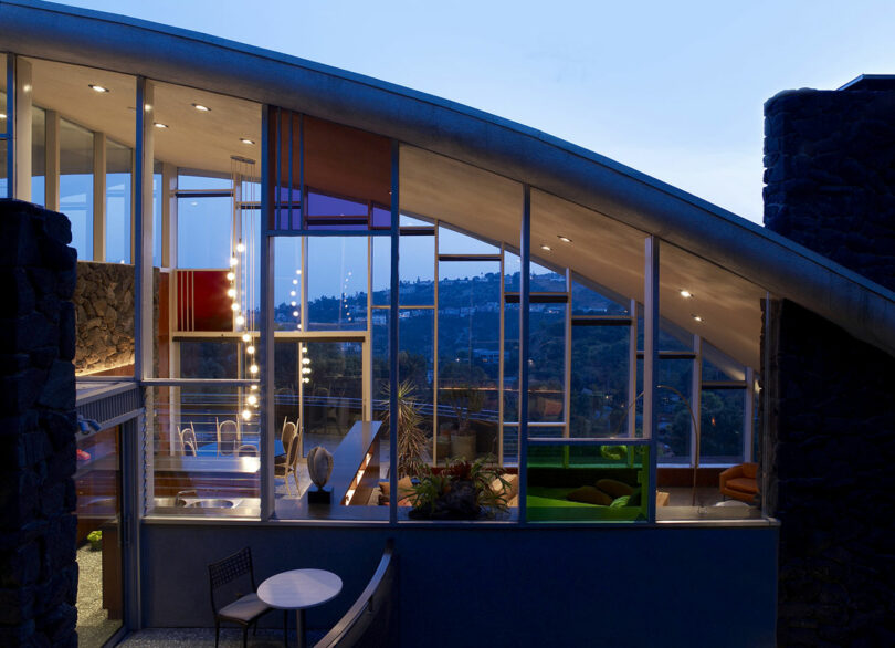 Modern glass-walled house at dusk, featuring a curved roof, interior lights, and visible furniture through large windows, overlooking a hilly landscape.