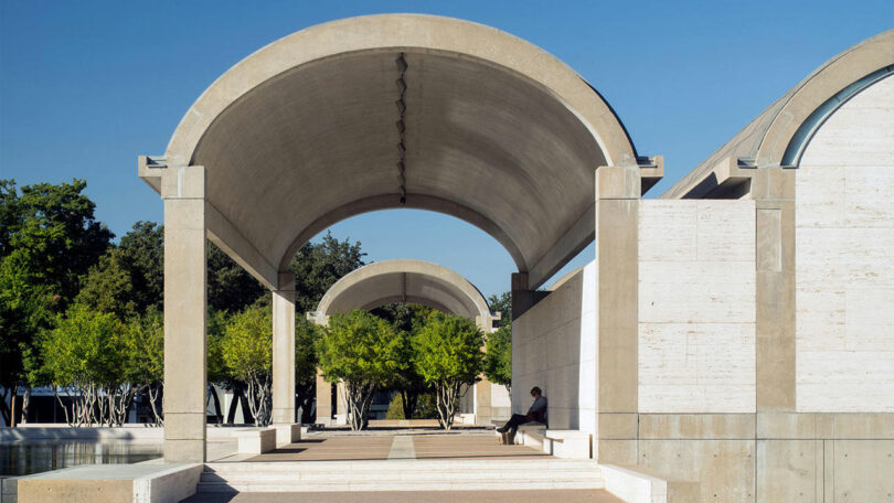 Concrete archways and columns with benches underneath, part of an outdoor architectural structure. Trees and clear blue sky in the background.