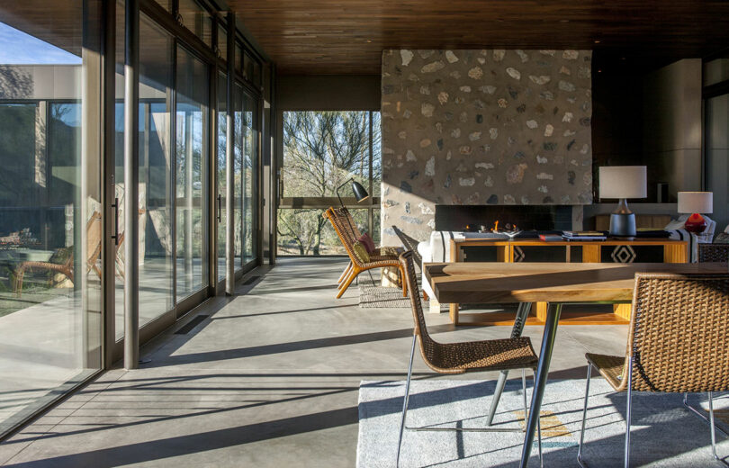 Modern interior with glass walls and stone accents. Woven chairs surround a wooden table on a rug. A desk and lamp are near a stone wall with trees visible outside.