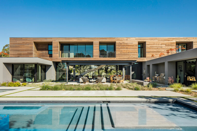 Modern two-story house with wood and glass facade, featuring an outdoor patio, garden, pool, and clear sky in the background.