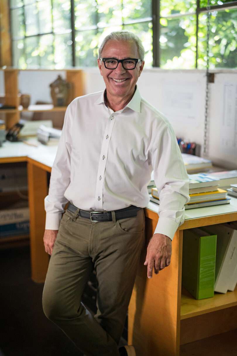 White-haired man in glasses and a white shirt leans against a desk in an office with shelves and a window behind him.