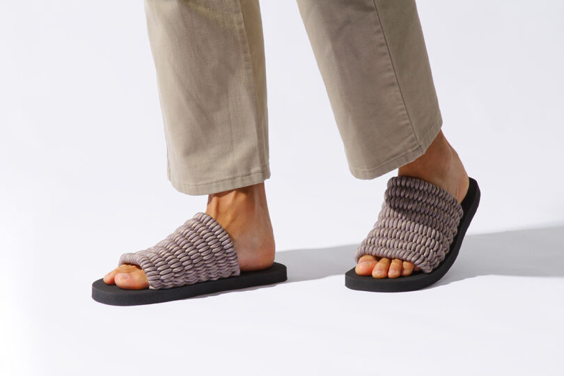 A person wearing beige pants and slip-on sandals with woven, textured straps. The sandals have black soles.