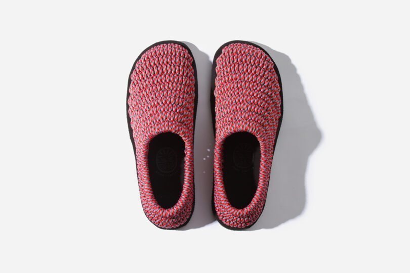 Top view of a pair of slip-on shoes with woven red, pink, and blue material, and black soles, placed on a white background.