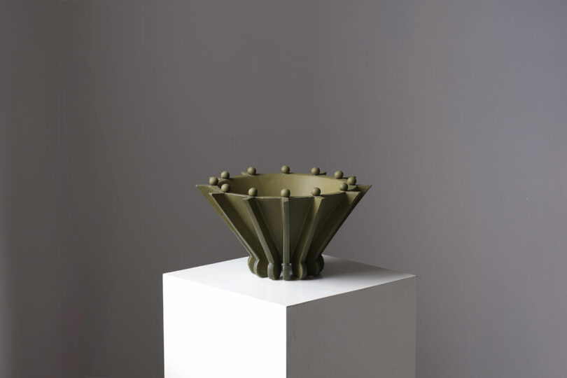 A green, geometric ceramic bowl with protruding knobs sits on a white pedestal against a gray background.