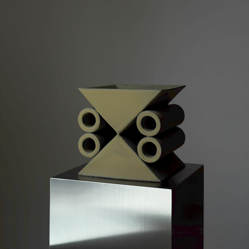 Geometric sculpture with triangular and cylindrical shapes on a metallic pedestal against a gray background.