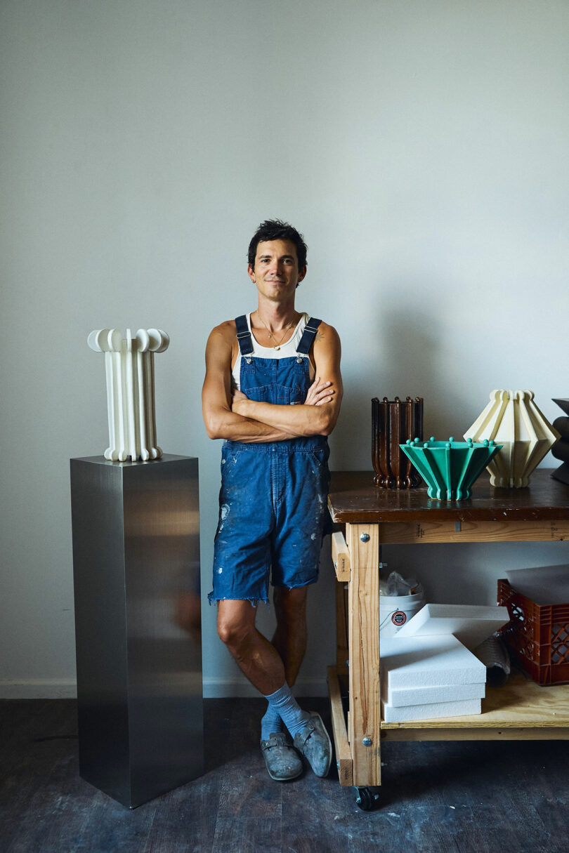 A person in blue overalls stands next to a display of sculptural objects on a pedestal and table in an art studio.