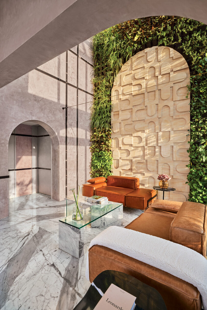 Modern living room with marble floor, brown leather sofas, glass coffee table, and a large green wall. An intricate wood panel design decorates one wall. Sunlight streams through an arched window.
