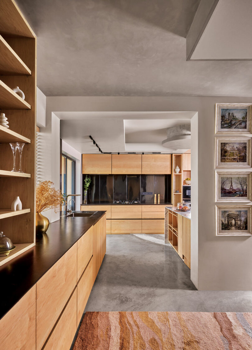 Modern kitchen with wooden cabinets, black countertops, and concrete floor. Shelves on the left display glasses and decor. Framed pictures hang on right wall.