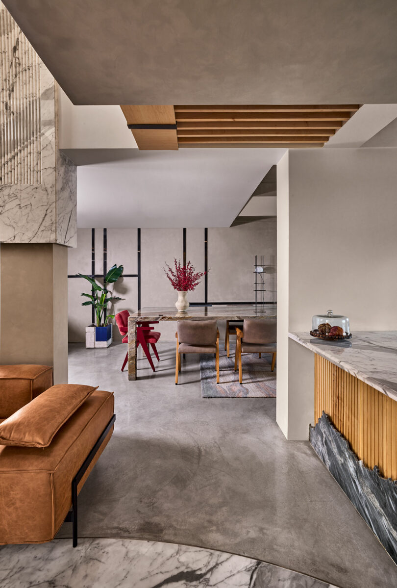 Modern living space with marble accents, leather seating, and a dining area with red chairs. A plant and a flower arrangement add color. The floor has a mix of concrete and decorative marble.