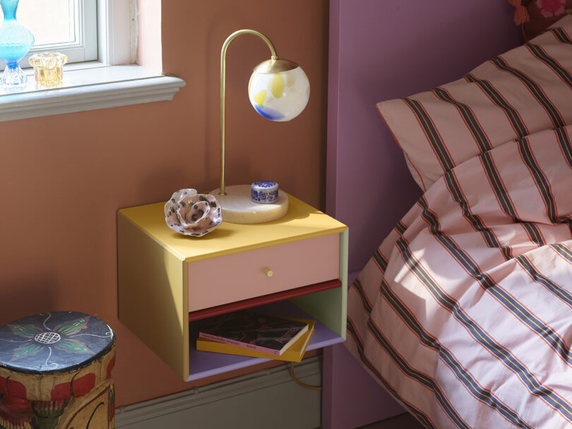 Bedside scene with a floating nightstand holding a globe lamp, circular decor, and a patterned book. Striped bedding and pastel walls with nearby window provide a cozy setting.