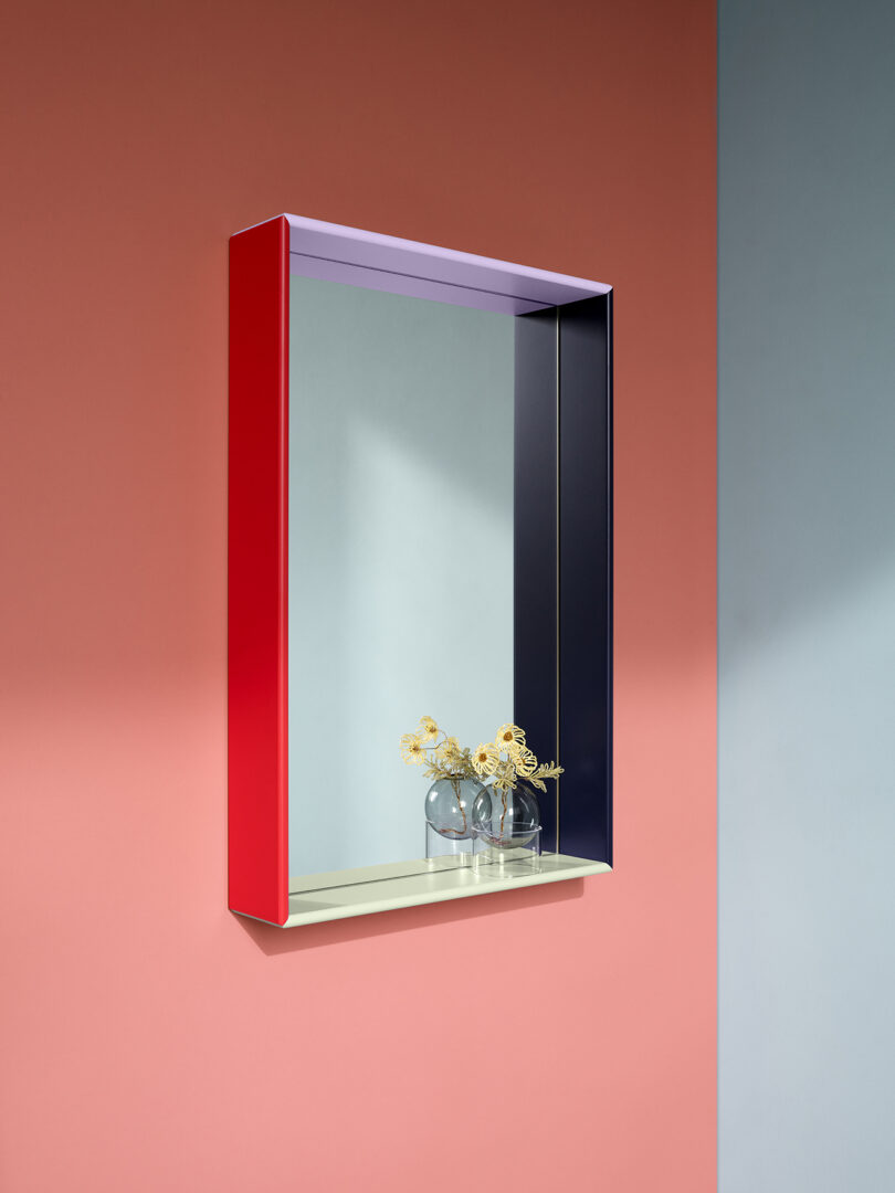 Rectangular mirror with a multicolored frame on a red wall. Two vases with yellow flowers are reflected in the mirror. Shadows cast due to directional lighting.