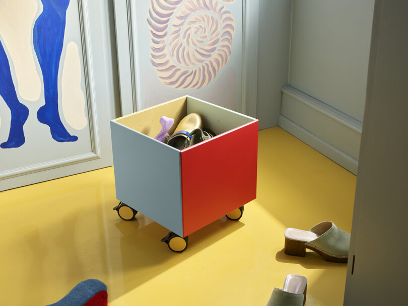 A colorful, wheeled storage cube contains shoes, placed on a yellow floor. Abstract artwork is visible on the wall.