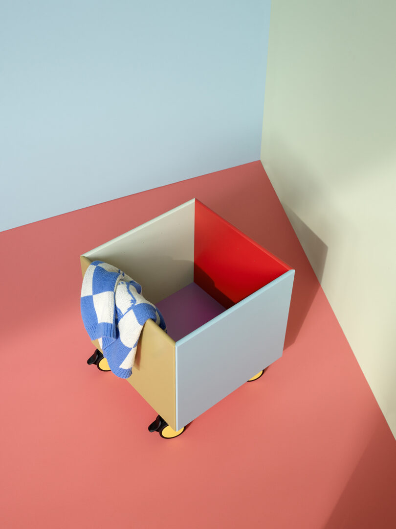 A multicolored storage box on wheels sits on a red surface, holding a blue and white checkered cloth over one side.