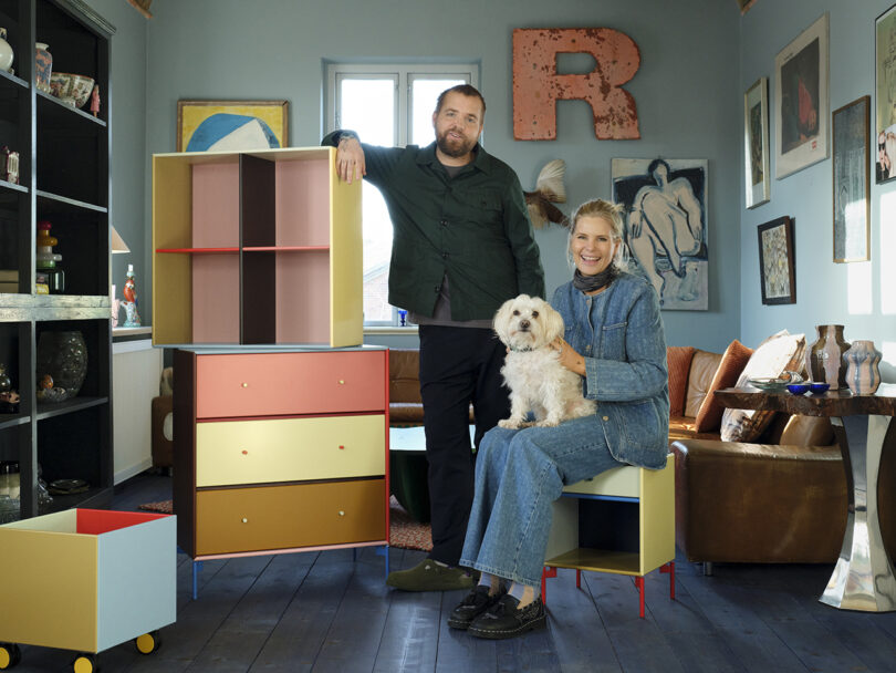 Two people are in a colorful room with eclectic decor. One sits with a small white dog on their lap, while the other stands beside geometric furniture pieces. Art and objects surround them.