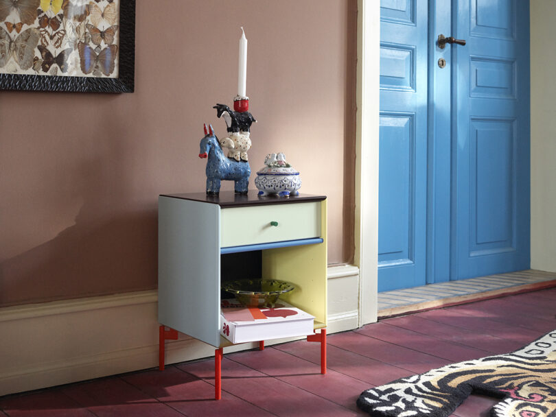 A small cabinet with a yellow and blue drawer, red legs, and ornaments on top stands against a peach wall. A blue door is in view on the right. A colorful rug covers the wooden floor.