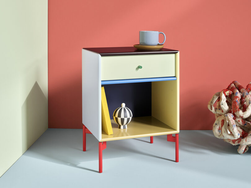 Modern side table with a drawer and open shelf, featuring a geometric ball and a yellow book. A ceramic sculpture and a mug sit nearby against a red and cream backdrop.