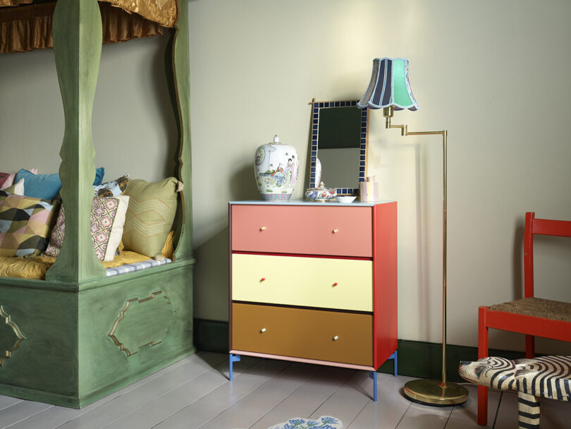 Room with a green bed, colorful dresser, mirrored lamp, and a red chair. Decor includes patterned pillows, a vase, and a standing lamp with a striped shade.