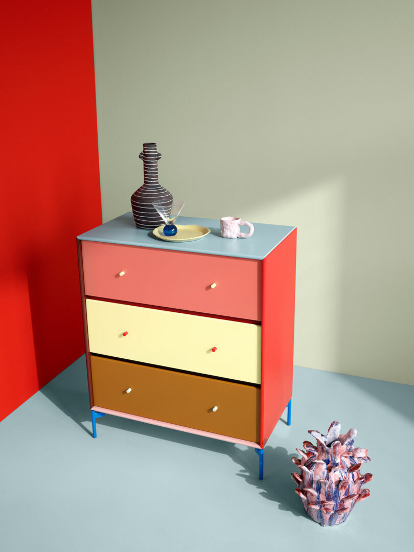 The KIMPOP Montana Furniture piece features a colorful chest of drawers with three vibrant drawers in red, yellow, and orange. On top, you'll find a vase, cup, and plate. A textured sculpture rests elegantly on the floor nearby.