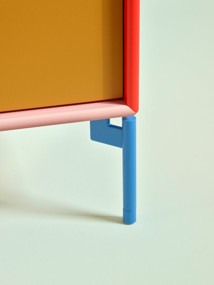 Close-up of KIMPOP modern furniture with a red frame, blue legs, and a yellow panel, set against a pale green background.