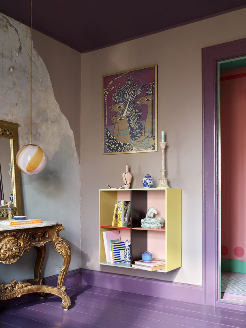 A room with purple floors, a decorative table, and a yellow bookshelf holding books and ornaments. A colorful light fixture hangs from the ceiling, and a framed abstract artwork is on the wall.