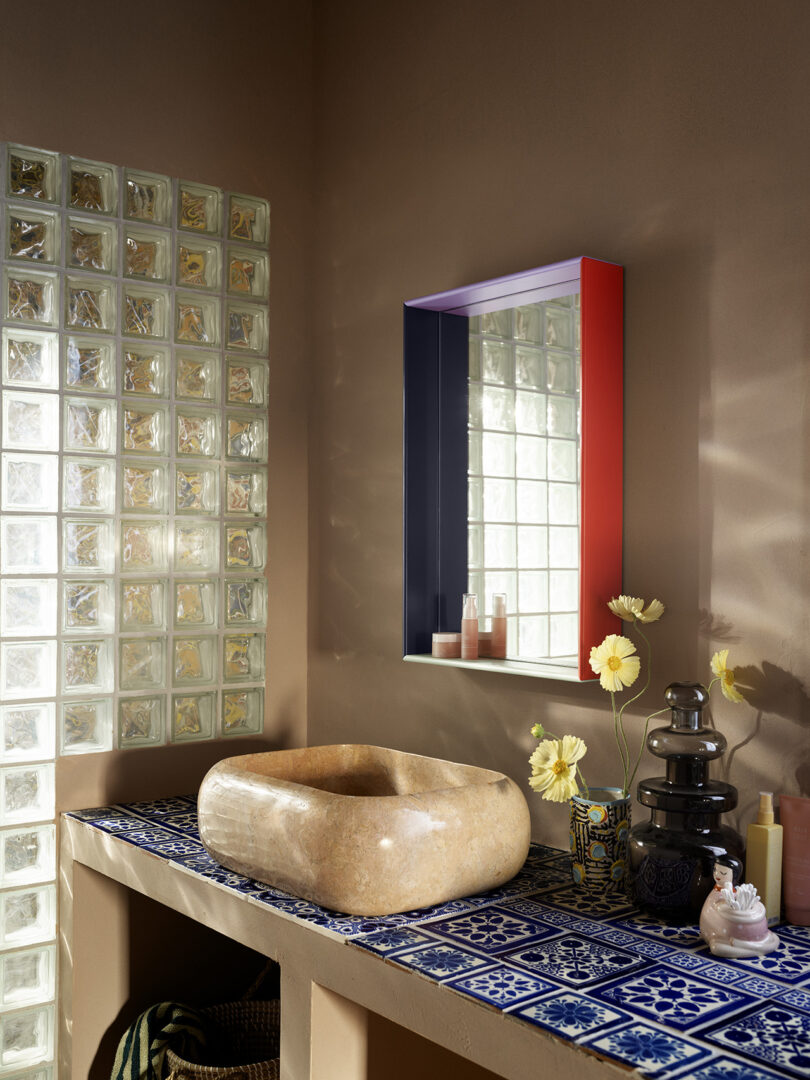 Bathroom with a stone sink on a blue tiled counter, yellow flowers, glass block wall, and a mirrored cabinet. Bottles and a textured vase are also on the counter.