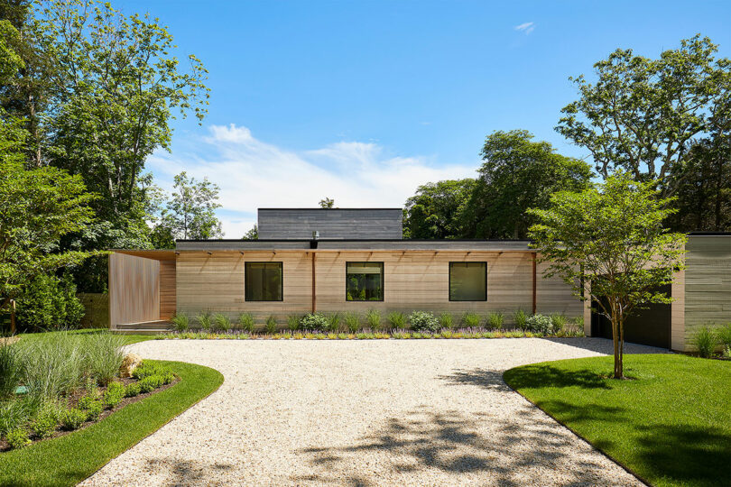 The Shift House is a modern single-story home with light wood siding, four windows, and a gravel driveway. It's gracefully nestled amid lush greenery and trees, all under a clear blue sky.