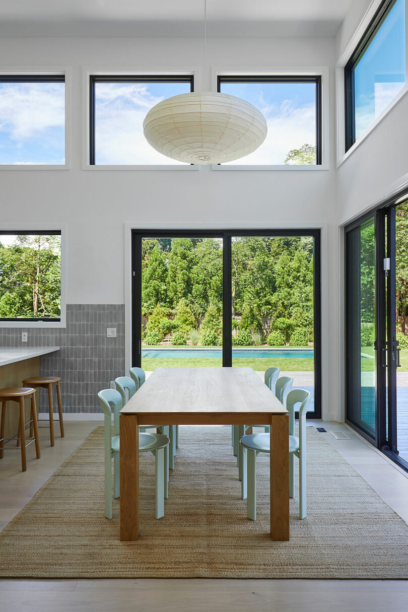 The Shift House offers a spacious dining area with a wooden table, light green chairs, a large round ceiling lamp, and expansive windows revealing a stunning view of the garden and pool.