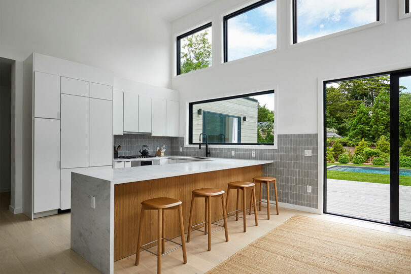 The Shift House kitchen boasts modern elegance with white cabinets and a marble island paired with wooden stools. Large windows illuminate the light wood flooring and gray tiled backsplash. A sliding glass door opens to a deck, inviting you to enjoy the greenery outside.