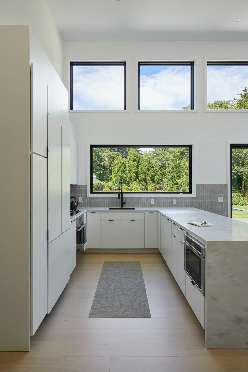 The Shift House features a modern kitchen with white cabinets, a marble countertop, and a light wood floor. Large windows flood the space with natural light, while a gray rug adds warmth. Green trees frame the view outside, seamlessly blending nature with contemporary design.
