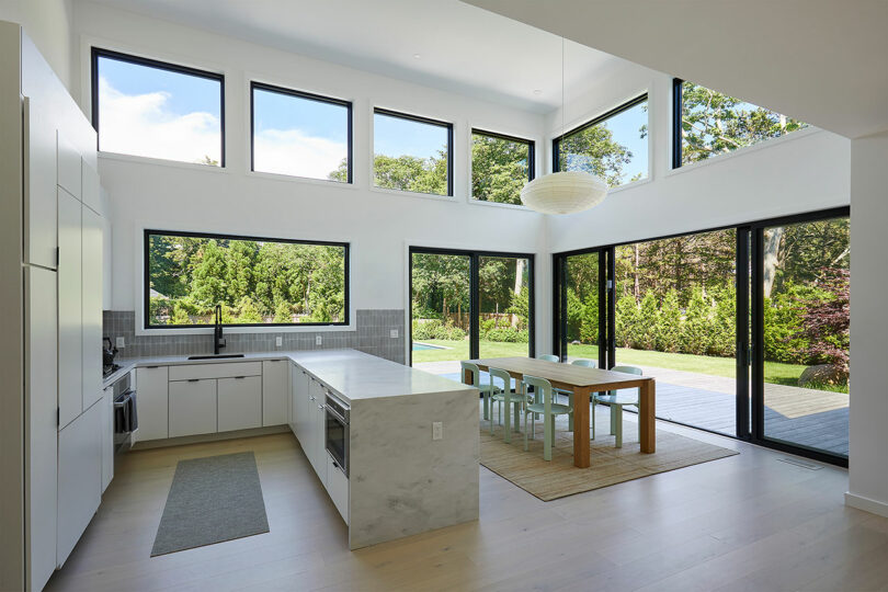 The Shift House features a modern kitchen and dining area with large windows, crisp white cabinets, an inviting island, and a wooden table. Glass doors seamlessly open to reveal the lush green garden beyond.